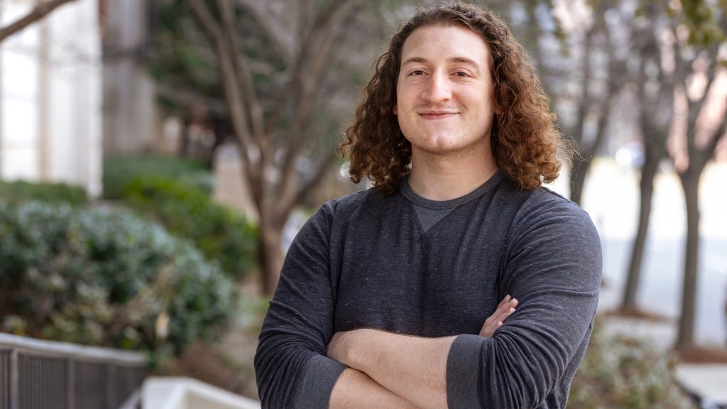 Young male student with shoulder length hair smiles directly at the camera standing outside on the Spalding campus