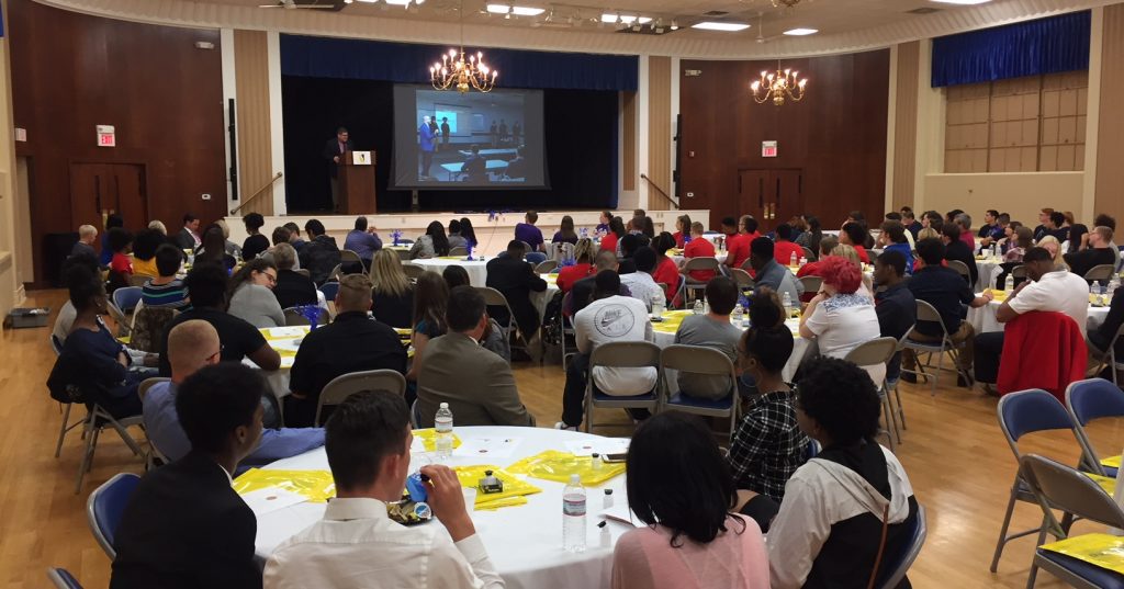Group of dozens of high school students and faculty seated at tables in a ball room, listening to remarks from Spalding Graduate Dean Kurt Jefferson speak from a podium on stage