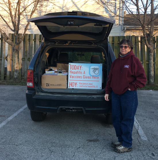 Nursing faculty Nancy Kern, standing in parking lot next to car with open hatchback, showing sign out the back that says 'Hepatitis A vaccines given here'