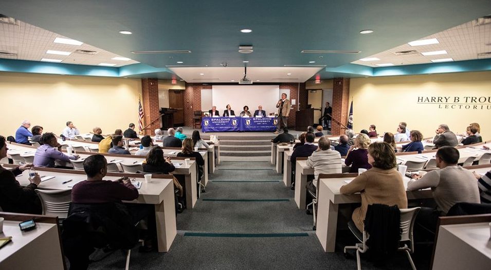 Panel of speakers sit at a table on stage as seated audience members look on at the Spalding ELC Troutman Lectorium