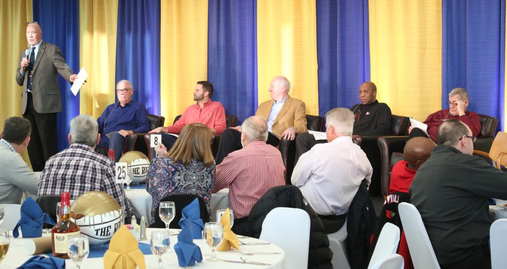 Tables of spectators watch Spalding AD Roger Burkman stand with a microphone on stage and address them and five other men seated in big chairs on stage: Mike Pratt, Luke Hancock, Dan Issel, Milt Wagner, Jerry Jones